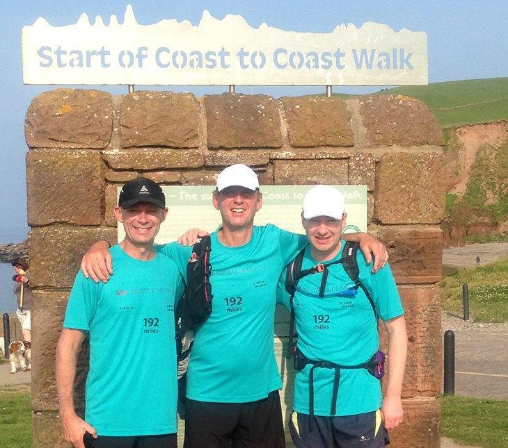 Three male fundraisers in blue t-shirts and caps standing in front of the start line of the Coast to Coast walk